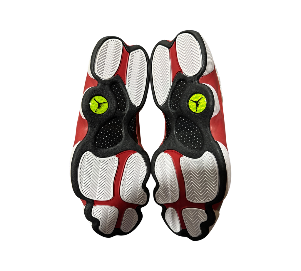 Genesis Boutique Jordan 13 Grey Toe sneakers showing the red, white, and black patterned rubber outsoles with logos.