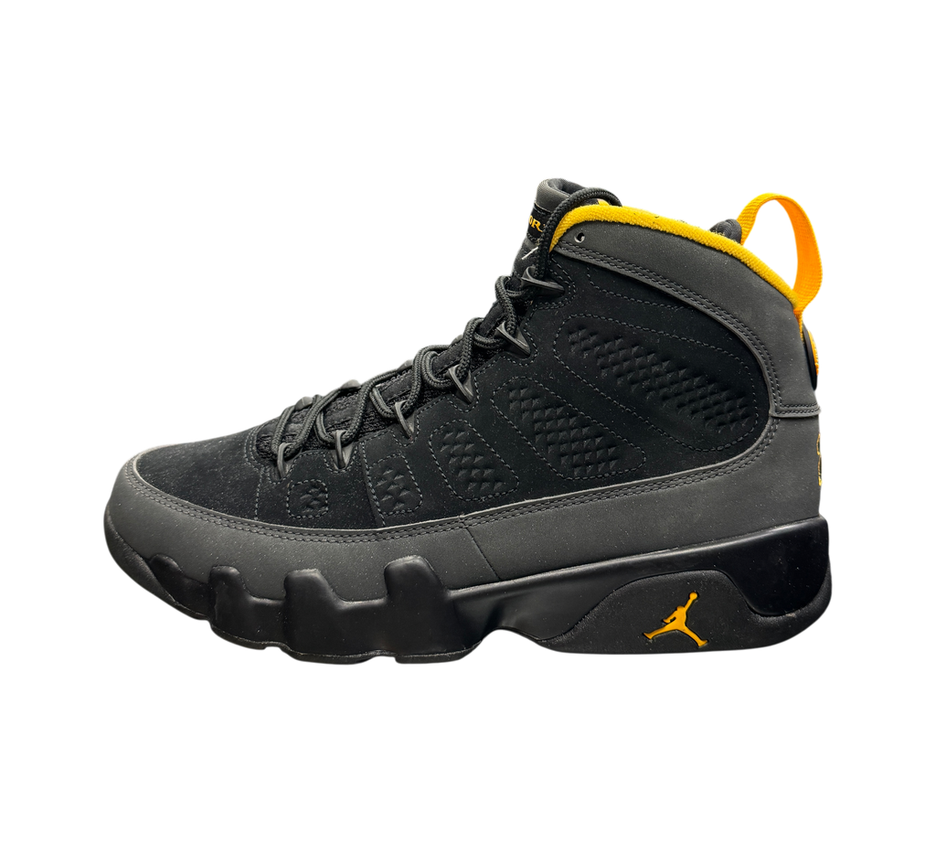 Genesis Boutique Jordan 9 Charcoal sneakers in black suede and nubuck with yellow accents on a glass display table.